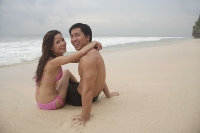 Couple sitting on beach, embracing, looking at camera - Asia Images Group