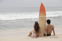 Couple sitting on beach, surfboard between them, looking out to sea - Asia Images Group