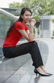 Young woman sitting on ledge outdoors, smiling at camera - Asia Images Group