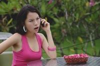 Young woman sitting at table outdoors, on the phone, angry expression on face - Asia Images Group