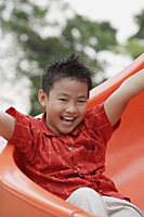 Boy coming down playground slide, arms outstretched, smiling - Asia Images Group