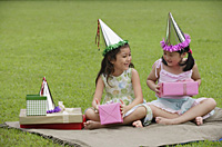 Two girls wearing party hats sitting on picnic blanket, holding pink gift boxes - Asia Images Group