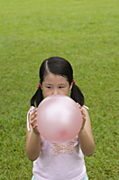 Girl standing on grass, blowing up pink balloon - Asia Images Group