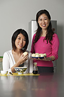 Mother and daughter in kitchen, younger woman sitting, older woman standing beside her holding a plate of Chinese buns - Asia Images Group