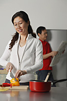 Mature woman in kitchen preparing a meal, man in background holding newspaper - Asia Images Group