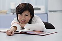 Woman leaning on table, book open in front of her, smiling at camera - Asia Images Group