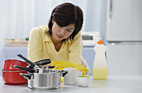 Woman in kitchen, leaning on kitchen counter, looking at stack of pots and pans - Asia Images Group
