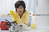 Woman leaning on kitchen counter, holding cleaning sponge, stack of pots and pans in front of her - Asia Images Group