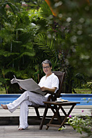 Mature man sitting by swimming pool, reading newspaper - Asia Images Group