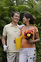 Couple in garden, woman holding bouquet of flowers and watering can, smiling at man next to her - Asia Images Group