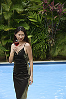 Woman standing and holding a glass of wine, swimming pool in the background - Asia Images Group