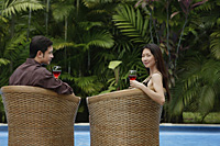 Couple sitting by swimming pool in rattan chairs, woman turning to look at camera - Asia Images Group