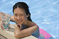 Woman leaning on edge of swimming pool, holding a drink, smiling at camera - Asia Images Group