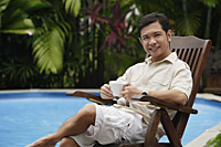 Man sitting by swimming pool, holding a cup of coffee - Asia Images Group