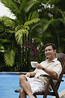 Man sitting by swimming pool, holding a cup of coffee, smiling - Asia Images Group
