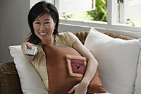 Woman sitting on sofa, holding TV remote control - Asia Images Group