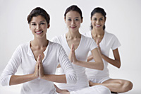 three women of mixed race sitting with legs crossed, arms in namaste, smiling at camera - Asia Images Group