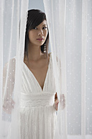woman in white dress, looking through curtains at camera - Asia Images Group