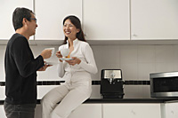 Woman sitting on counter in kitchen with man standing next to her, looking at each other, holding coffee cups and smiling - Asia Images Group