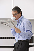Man in kitchen getting ready for work, drinking coffee and reading newspaper, tie loose around neck - Asia Images Group