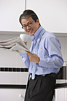 Man in kitchen getting ready for work, drinking coffee and reading newspaper, smiling at camera with tie loose around neck - Asia Images Group