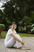 Woman sitting on porch in tropical setting, arms wrapped around legs, looking at camera, smiling - Asia Images Group
