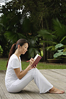 side profile of Woman sitting on porch in tropical setting, writing in diary, journal. - Asia Images Group