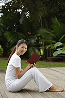 side profile of woman sitting on porch in tropical setting, writing in journal, diary, and looking at camera, smiling - Asia Images Group