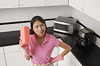 woman standing in kitchen, wearing gloves and apron for cleaning, holding sponge and thinking. - Asia Images Group