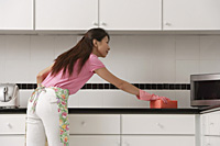 woman standing in kitchen, wearing gloves and apron and cleaning counter with big pink sponge. - Asia Images Group