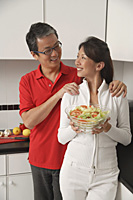 Man and woman in kitchen holding salad bowl and looking at each other smiling - Asia Images Group