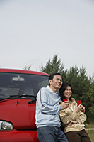 man and woman leaning against front of red van, holding drinks, nature, camping, outdoors - Asia Images Group