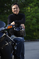 Man leaning on motorcycle, holding helmet, smiling at camera - Asia Images Group