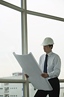 man wearing hard hat, holding blue prints, looking out window - Asia Images Group