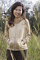 Woman standing in tall grass, hands in pocket, smiling at camera, nature - Asia Images Group