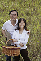 Man and woman standing outdoors in nature, holding picnic basket, smiling at camera - Asia Images Group