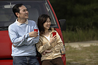 Man and woman leaning against red van, drinks in hand, camping, outdoors - Asia Images Group