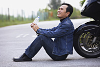 Man leaning against motorcycle, sitting on road drinking water - Asia Images Group