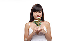 Young woman wearing white dress, holding small plant and smelling flower - Asia Images Group