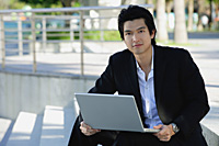 A man uses his laptop outdoors while he looks at the camera - Asia Images Group