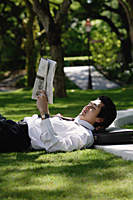 A man lies down and reads the newspaper in the park - Asia Images Group