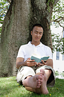 A man leans against a tree and reads in the park - Asia Images Group