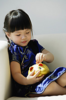 A small girl in blue silk cheongsam plays with a piggy bank - Asia Images Group