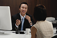 A man sits at his desk while he talks with a woman at work - Asia Images Group