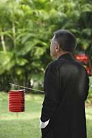 Man in traditional clothing, standing in park holding Chinese lantern - Asia Images Group
