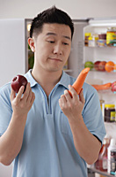 Man holding an apple and a carrot, looking frustrated - Asia Images Group