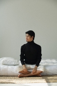 Young man sitting on bed, looking sideways - Asia Images Group