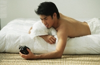 Young man lying in bed, holding alarm clock - Asia Images Group