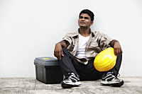 man sitting on ground holding construction hat - Asia Images Group