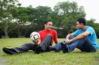 Two men sitting on grass, talking, one holding soccer ball - Asia Images Group